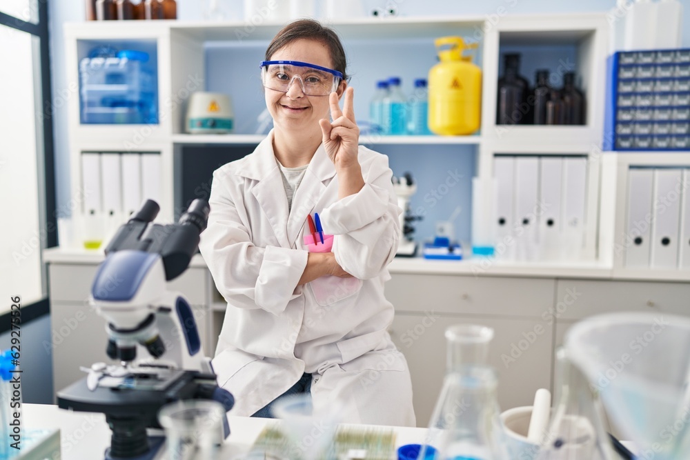 Hispanic girl with down syndrome working at scientist laboratory smiling with happy face winking at the camera doing victory sign with fingers. number two. Hispanic girl with down syndrome working at scientist laboratory smiling with happy face winking at the camera doing victory sign with fingers. number two.