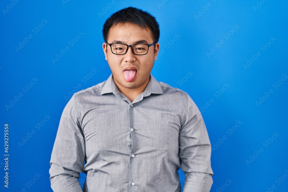 Young chinese man standing over blue background sticking tongue out happy with funny expression. emotion concept. Young chinese man standing over blue background sticking tongue out happy with funny expression. emotion concept.