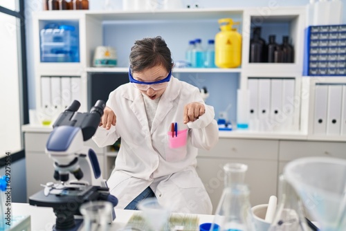 Hispanic girl with down syndrome working at scientist laboratory pointing down with fingers showing advertisement  surprised face and open mouth