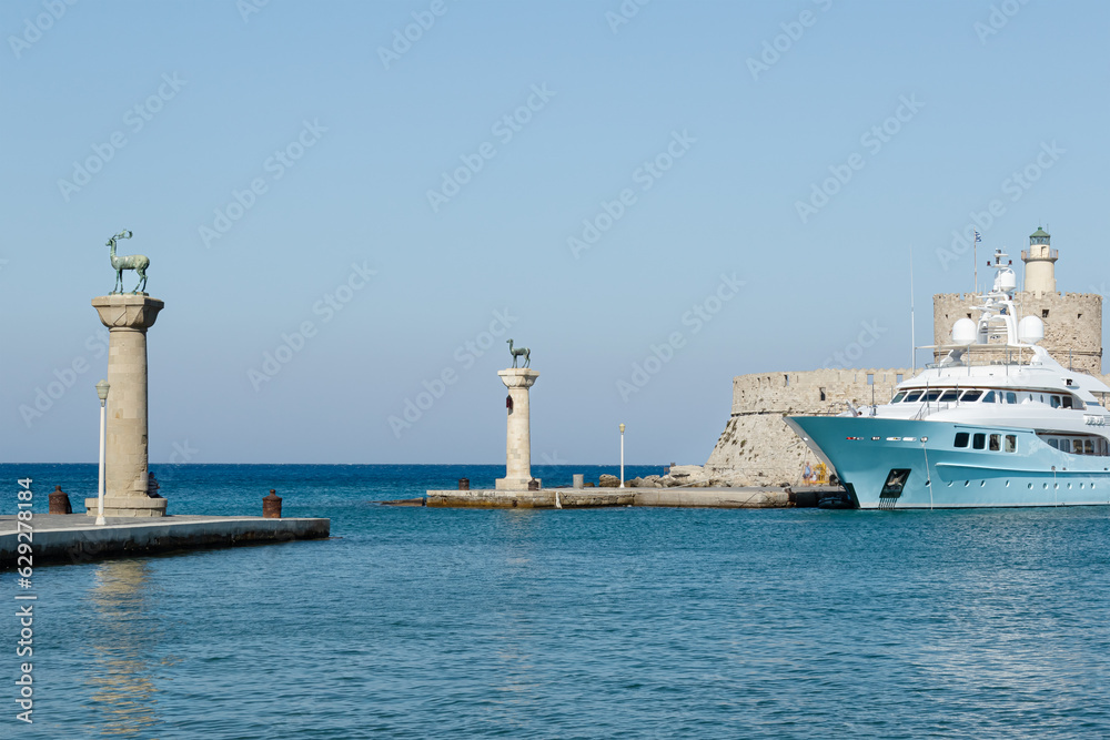 selective focus on columns of the Colossus of Rhodes and Fortress St. Nicholas with  tourist boat in front of it, old harbor entrance