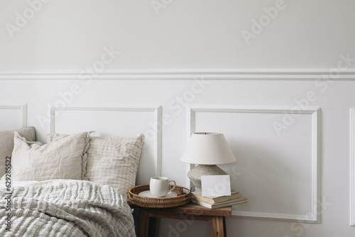 Blank greeting card, invitation mockup. Breakfast in bed concept. Cup of coffee, table lamp with linen shade, vintage wooden night stand. Linen beddings, elegant classic bedroom interior, white wall.
