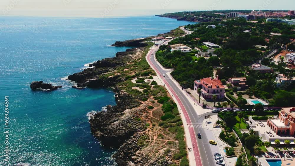 Drone point of view road leading along rocky coastline in the touristic resort of Cascais coastal town. Atlantic Ocean view and townscape, aerial view. Portugal. Travel destinations concept