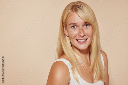Portrait of a young beautiful woman with freckles smiling and looking at the camera against a beige background