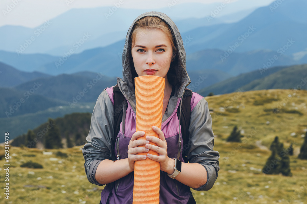 Fototapeta premium A tourist girl with a mat in her hands, dressed in a windbreaker with a hood, looks into the camera against the backdrop of mountain peaks.