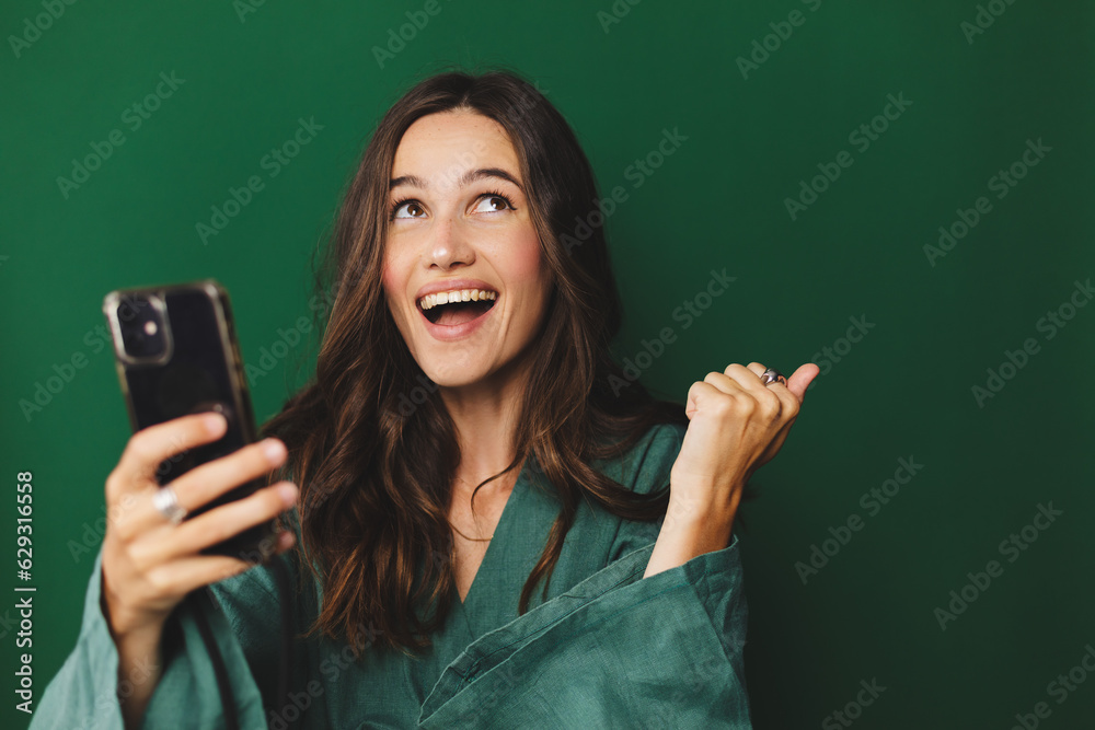 Fun young woman hold in hand use mobile cell phone doing winner gesture clenching fists isolated on green background studio. Girl using smartphone apps winning online celebrating discount gift voucher - obrazy, fototapety, plakaty 