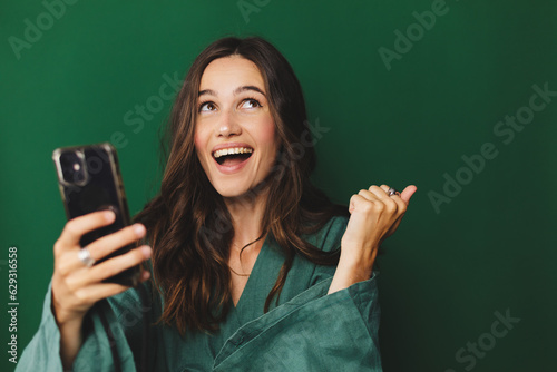 Fototapeta Fun young woman hold in hand use mobile cell phone doing winner gesture clenching fists isolated on green background studio