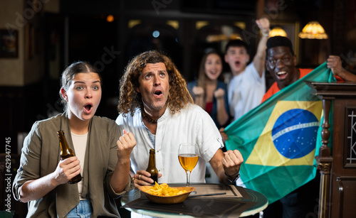 Excited interested young adult couple watching match at table in sports bar, rooting for favorite team against background of fans with national flag of Brazil..