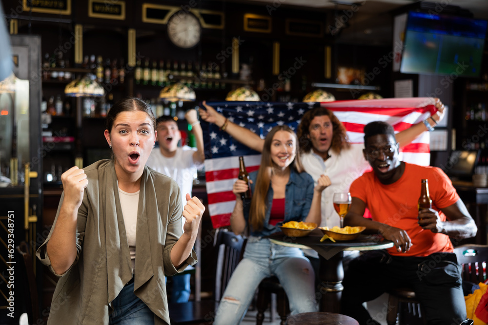 Cheerful diverse soccer supporters with flag of USA spending time together with pint of beer and snacks in the pub