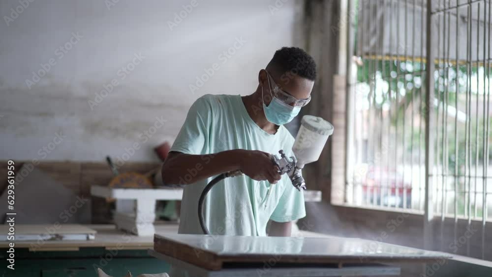 One young black worker applying paint spray on wooden surface at carpentry workshop One young black worker applying paint spray on wooden surface at carpentry workshop