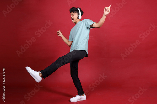 Full length attractive Asian young man listening music in headphones singing and dancing isolated vibrant red color background © Queenmoonlite Studio