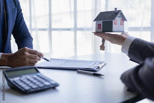 Female real estate agent handing house keys to client with small mockup of house and signing contract on papers on table in office close up pictures