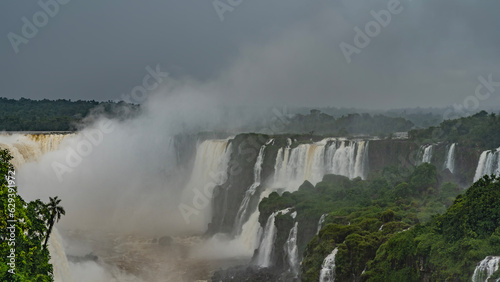 Beautiful cascades of waterfalls collapse into a stormy river. Spray and fog rise into the sky. Green tropical vegetation on ledges. Iguazu Falls. Brazil.