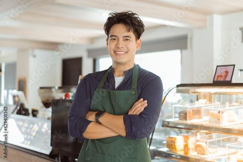 Young Asian male barista in apron having arms folded and smiling while looking at camera with copy space