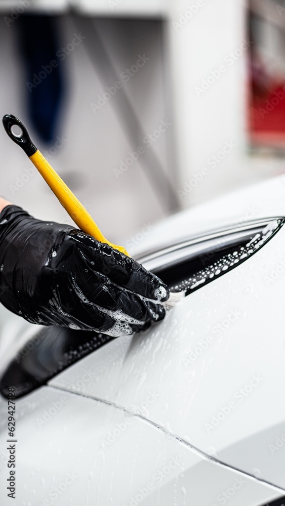 Employee of a car detailing studio or car wash carefully cleans the area around the front headlight with a brush in a white car