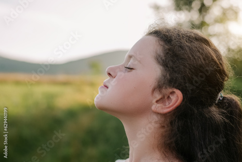 Closeup of relaxed little child with close eyes in outdoors breathe fresh air from nature during warm sunset. Portrait beautiful kid girl enjoying green park for good health. Childhood concept