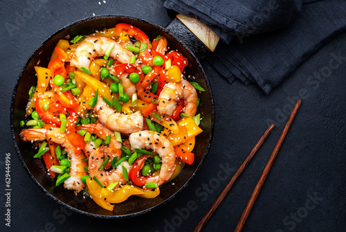 Stir fry shrimps with  red paprika, green peas, chives and sesame seeds in frying pan. Asian cuisine dish. Black kitchen table background, top view