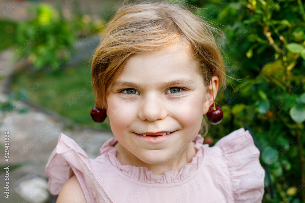 Little preschool girl picking and eating ripe cherries from tree in garden. Happy child with glasses holding fresh fruits. Healthy organic berry cherry summer harvest season. With cherry as earrings