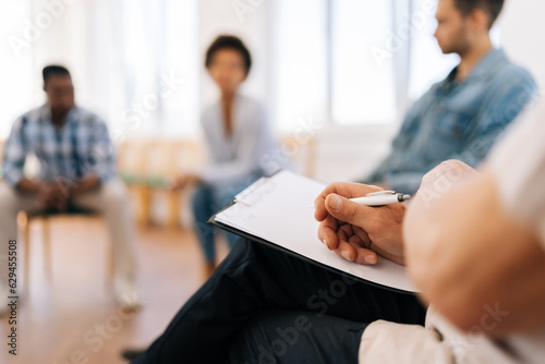 Close-up selective focus shot of unrecognizable mature male psychologist with clipboard having group therapy session with diverse male and female patient sitting in circle. Concept of mental health.
