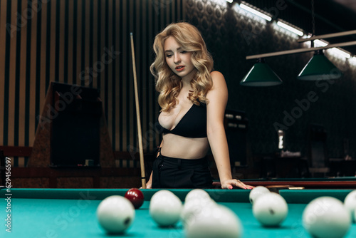 Blond woman playing enjoying billiard, hold cue, white billiard balls on table with green surface in billiard club. Pool game snooker pyramid player
