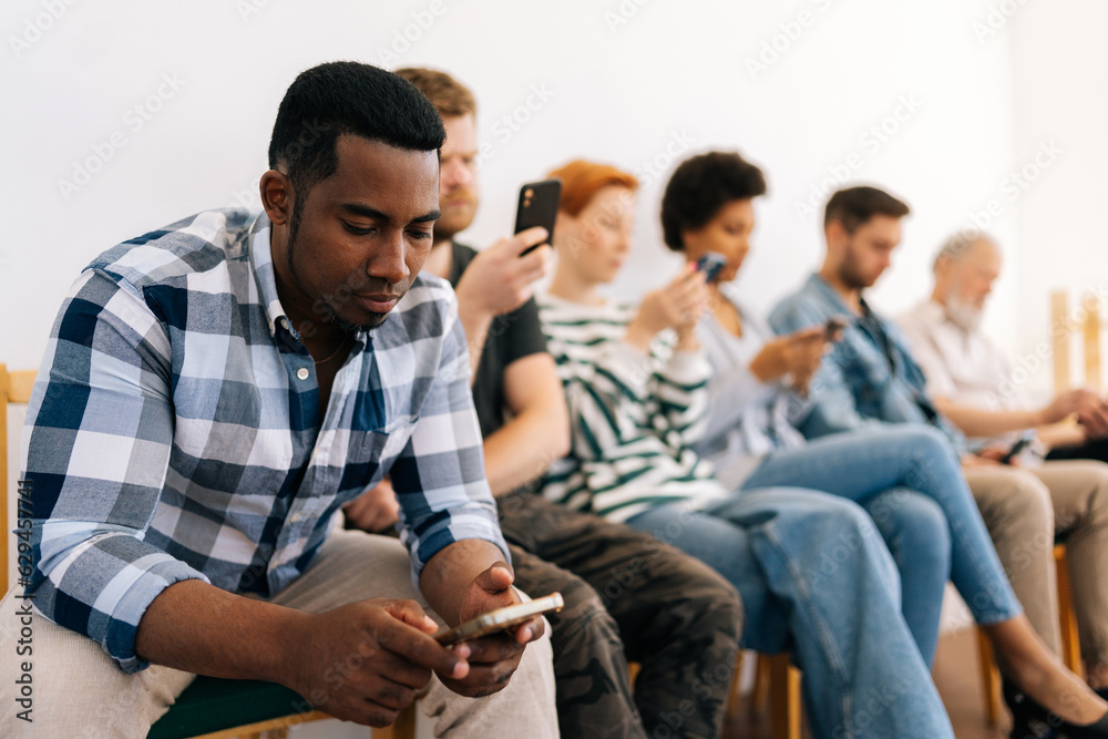 Selective focus of bored multicultural and different ages people in casual clothes sit on chairs in row waiting for job interview using typing mobile phones, recruitment hiring hr concept. Selective focus of bored multicultural and different ages people in casual clothes sit on chairs in row waiting for job interview using typing mobile phones, recruitment hiring hr concept.