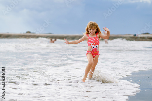 Happy Child, Little Preschool Girl in Swimmsuit Running And Jumping In The Waves During Summer Vacation On Exotic Tropical Beach. Family Journey On Ocean Coast.