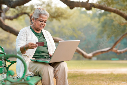 Indian senior woman using laptop and bank card at park.