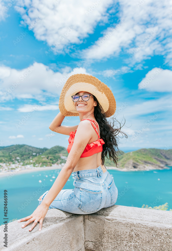 Portrait of tourist girl in a hat at a viewpoint with a beach in the background. Happy tourist female sitting at a viewpoint with beach in the background
