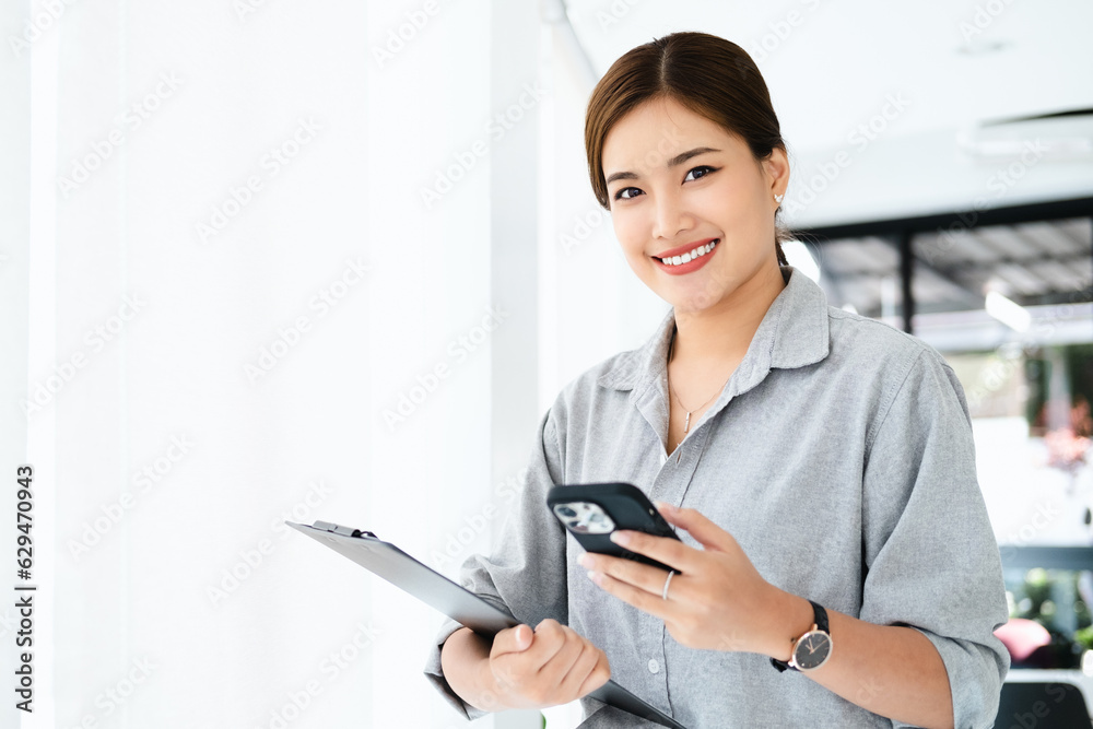 Young asian business woman wearing shirt using tablet and phone standing in modern workspace. Smiling confident business leader looking at camera and standing in an office. Young asian business woman wearing shirt using tablet and phone standing in modern workspace. Smiling confident business leader looking at camera and standing in an office.