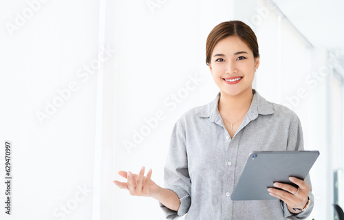 Young asian business woman wearing shirt using tablet standing in modern workspace. Smiling confident business leader looking at camera and standing in an office.