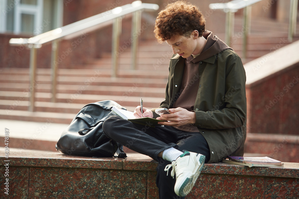 Writing by using pen. Young student with curly hair is outdoors near university Writing by using pen. Young student with curly hair is outdoors near university