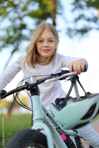 Cheerful girl riding her bike in a green park with a pink water bottle. A cute kid enjoying her summer day on a bicycle with a helmet for safety. © Andrii Zastrozhnov