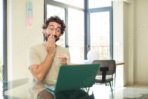young adult bearded man with a laptop laughing at you, pointing to camera and making fun of or mocking you