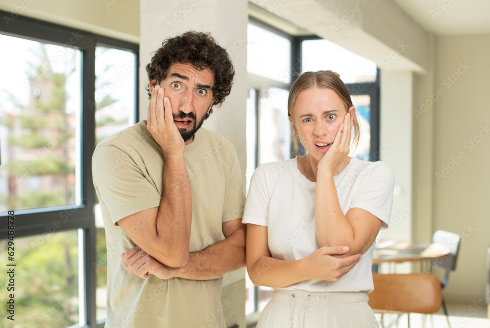 young adult couple open-mouthed in shock and disbelief, with hand on cheek and arm crossed, feeling stupefied and amazed young adult couple open-mouthed in shock and disbelief, with hand on cheek and arm crossed, feeling stupefied and amazed