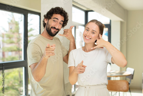 Fototapeta Naklejka Na Ścianę i Meble -  young adult couple smiling cheerfully and pointing to camera while making a call you later gesture, talking on phone