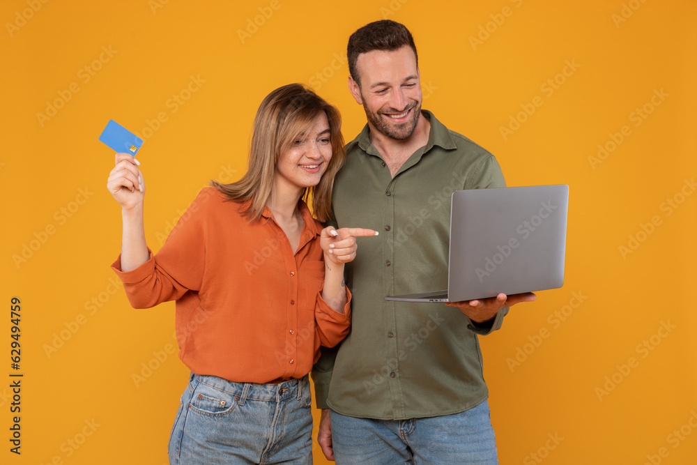 Happy young couple in casual holding modern pc laptop and plastic bank card, lady pointing at computer screen, spouses shopping online or booking tickets, yellow background