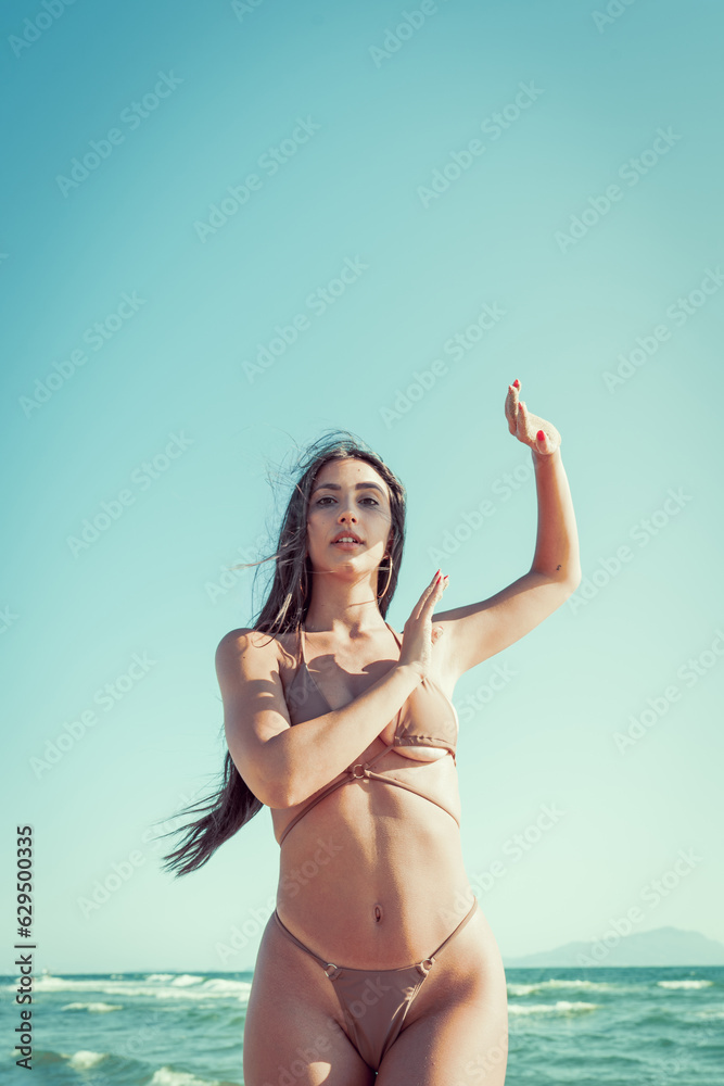 Photo shoot of a beautiful young woman posing wearing sea costume on the sand near the ocean for holidays with light blue sky as background in a sunny day