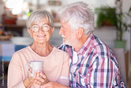 Senior white-haired man embracing and looking with affection at his wife drinking a cup of coffee or tea. Forever love, elderly couple in love