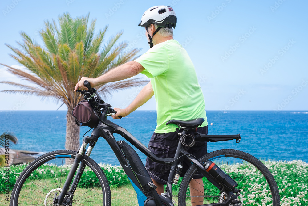 Elderly athletic man with electric bicycle in outdoors excursion at sea. Senior man in helmet enjoying freedom and healthy lifestyle looking at horizon over water