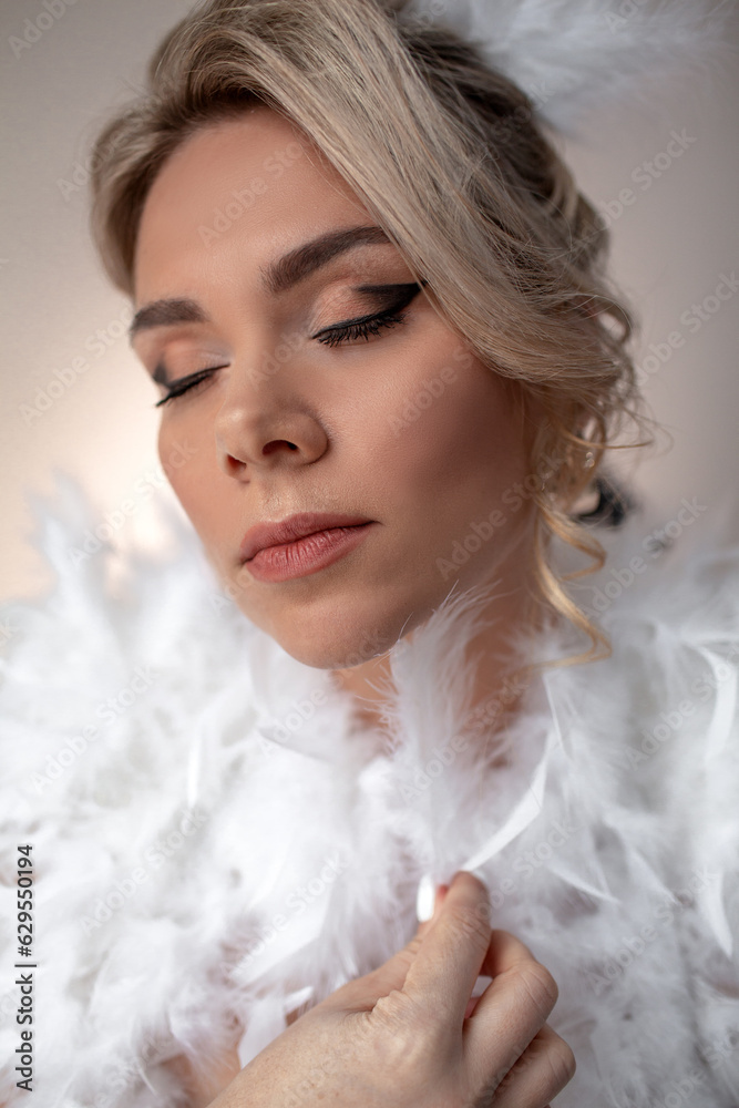 Portrait of a beautiful bride with a wedding hairstyle dressed in a white dress with feathers. A girl with a professional makeup against the wall