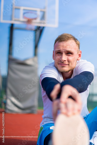 A tall guy basketball player sits on a basketball court outside and stretches his muscles before the start of practice close-up 