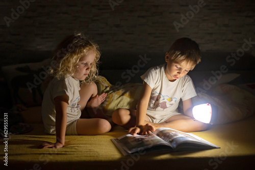 Two blond cute childrem, boy and girl, siblings, lying under the cover in bed, reading book together with small light photo