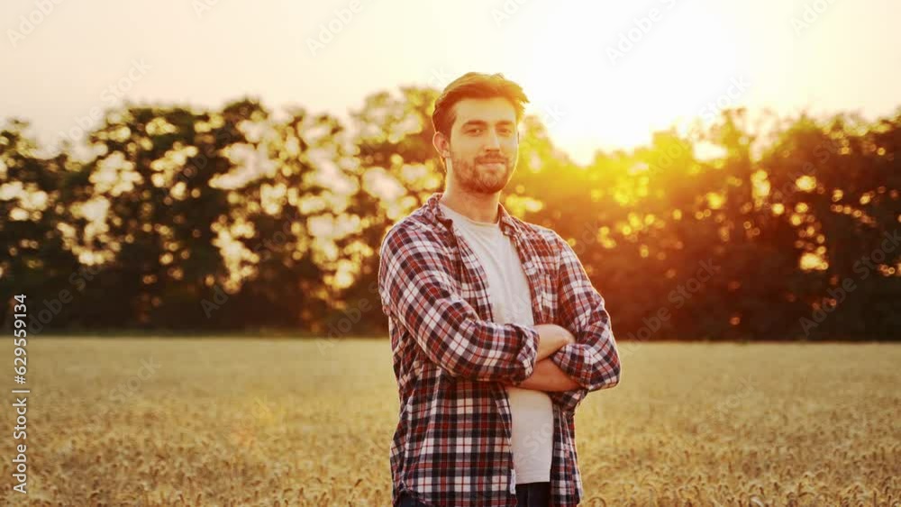 Portrait of farmer standing in ripe wheat field with arms crossed on chest. Proud agronomist looking at camera on a farmland on sunset. Rich harvest of cultivated cereal hybrid crop. Harvesting season Portrait of farmer standing in ripe wheat field with arms crossed on chest. Proud agronomist looking at camera on a farmland on sunset. Rich harvest of cultivated cereal hybrid crop. Harvesting season