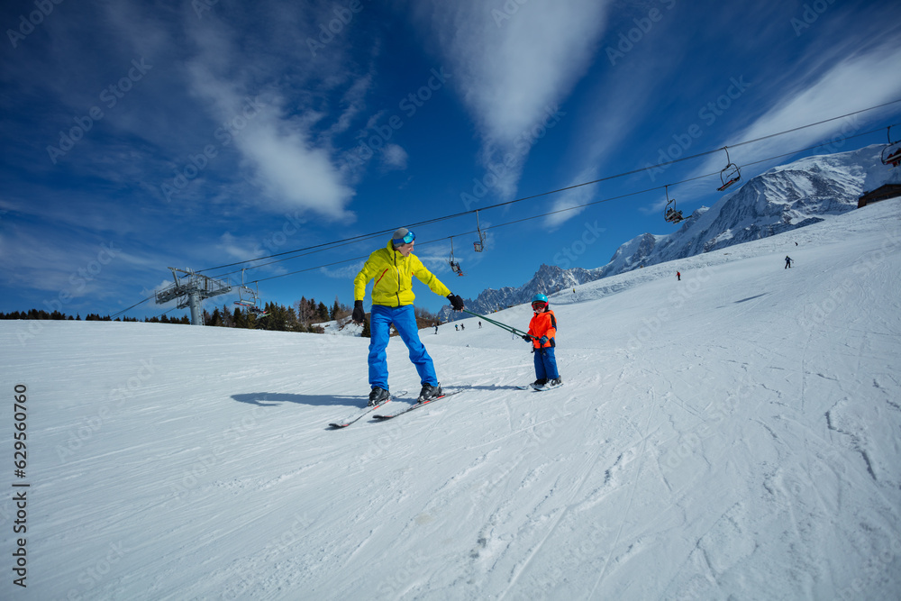 Father go downhill teach child to ski connected by poles