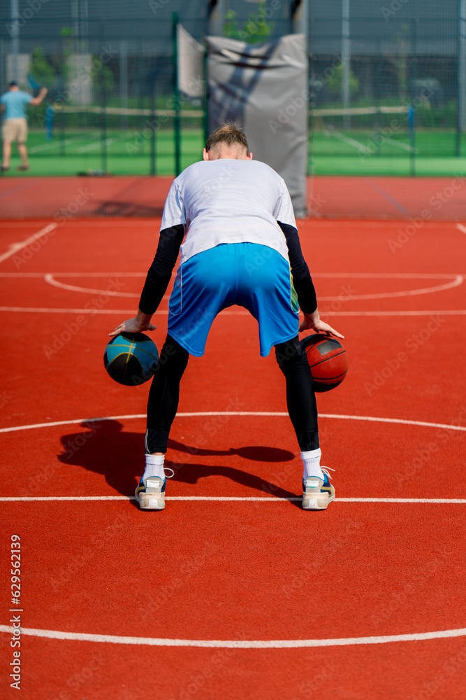 A tall guy basketball player with two balls shows off his dribbling skills while practicing on a basketball court in the park 