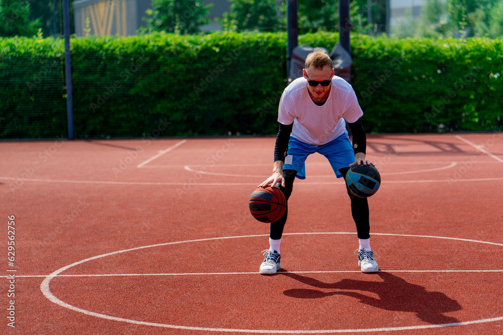 Obraz premium A tall guy basketball player with two balls shows off his dribbling skills while practicing on a basketball court in the park 
