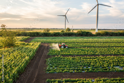 Aerial view of crop sprayer spraying pesticide or herbicides field at sunset. Protection plants to increase crop yield. Blue sky and windmill turbines on background. Bio food and green energy together