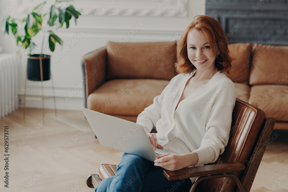 Redhead freelancer joyfully works from home, using a modern laptop for remote tasks, relishing online communication while awaiting responses.