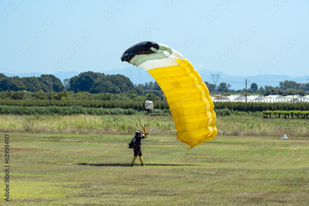 Stunning view of a skydiver with a yellow parachute landing on a green lawn during a sunny day.