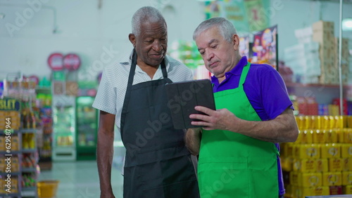 Two senior diverse employees working at supermarket looking at tablet device, teamwork scene of older manager orienting employee at grocery store, job occupation concept