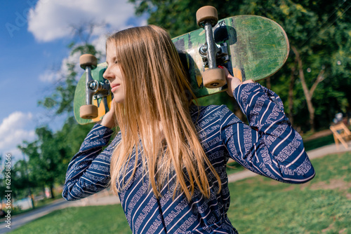 Portrait of a young smiling skater girl with long hair holding a skateboard on her shoulders against a background of trees and sky